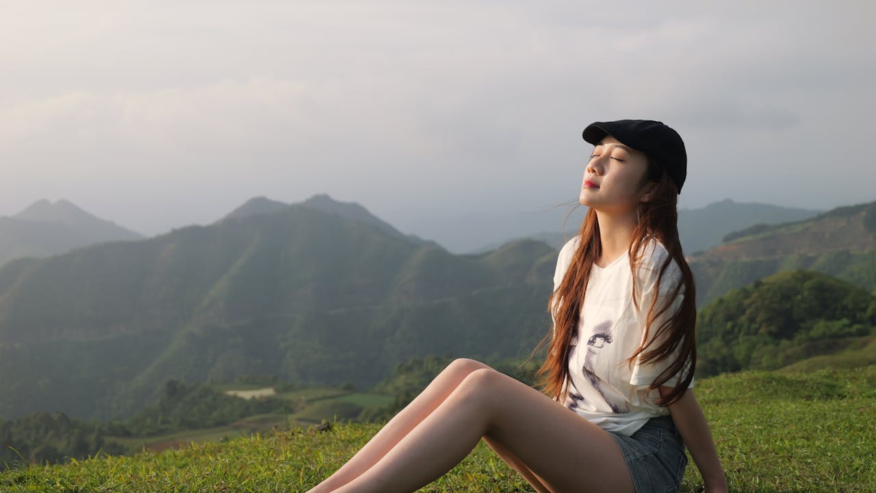 Woman enjoying tranquil moment on a grassy hilltop with mountains in the background.
